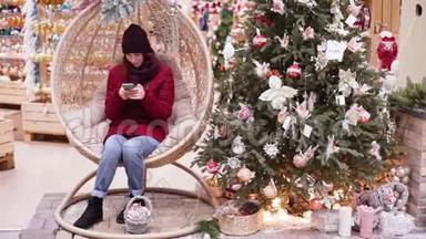一个年轻漂亮的女人在圣诞商店，使用智能手机。 女人在商店里走来走去挑选圣诞装饰品